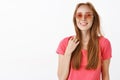 Studio shot of cute happy and emotive young redhead female in pink sunglasses and t-shirt touching strand of hair and Royalty Free Stock Photo