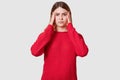 Studio shot of displeased female suffers from headache, dressed in red sweater, keeps hands on temples, has upset facial