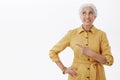 Studio shot of dreamy and energized kind cute elderly woman in yellow stylish trench coat pointing and looking at upper