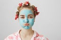 Studio shot of funny housewife in hair-curlers and nightwear, sulking while standing with facial mask and doing