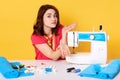 Studio shot of girl seamstress sitting at white desk with sewing machine, spreding hands, does not know how to switch it.