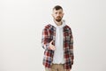 Studio shot of gloomy unconfident european guy with fair hair in trendy plaid shirt over hoodie, pulling hand towards