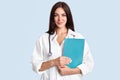 Studio shot of good looking female doctor wears white robe, has phonedoscope on neck, holds folder with medical documents, looks d Royalty Free Stock Photo