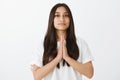 Studio shot of grinning good-looking female coworker in white t-shirt, holding hands in pray and smiling with pleased