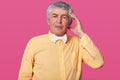 Studio shot of handsome old man keeps hand on temple, has headache, wears yellow shirt and white bow tie, has embrassed facial Royalty Free Stock Photo