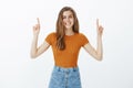 Studio shot of happy charming fair-haired woman in trendy outift, raising index fingers and pointing up while smiling