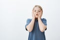 Studio shot of impressed amazed cute male child in blue t-shirt, holding palms on cheeks, holding breath and gasping