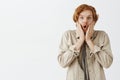 Studio shot of impressed excited happy redhead male with wavy hair and beard in outdoor clothes holding hands on face