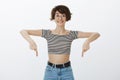 Studio shot of impressed happy and excited woman in round eyewear and striped cropped top, pointing down with index