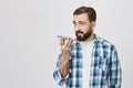 Studio shot of male european adult holding phone near mouth while talking to it on speaker, standing against gray Royalty Free Stock Photo