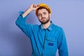 Studio shot of man wearing blue uniform and yellow building helmet looking at camera, keeping hand on his hard hat, handsome guy Royalty Free Stock Photo