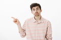 Studio shot of miserable upset nerdy guy with beard and moustache, pointing left with index finger, making unhappy smile