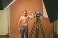 Muscular Indian man with mustache shirtless while vlogging against brown background