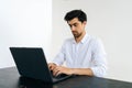 Studio shot of pensive young Caucasian man working online on laptop computer sitting at table in room with light Royalty Free Stock Photo