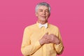 Studio shot of pleasant looking kind hearted eldery man keeps hands on chest, expresses gratitude, dressed in yellow shirt in ione Royalty Free Stock Photo