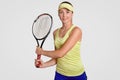 Studio shot of pleasant looking sporty determined female wears court cap, t shirt and shorts, holds tennis racquet, being active p