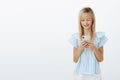 Studio shot of pleased good-looking small female kid with blond hair in blue blouse, holding smartphone and smiling at