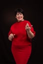 Studio shot of a senior adult smiling woman wearing a red dress on a dark background. She is smiling cheerfully gesticulating