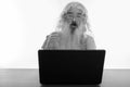 Studio shot of senior bearded man looking shocked while using laptop on wooden table Royalty Free Stock Photo