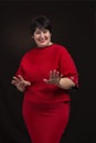 Studio shot of a senior woman wearing a red dress on a dark background. She is smiling cheerfully gesticulating