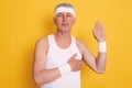 Studio shot of serious concentrated mature man posing against yellow wall, wearing white sleeveless t shirt and head band, showing Royalty Free Stock Photo