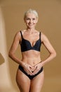 Studio shot of smiling mature blonde woman in black underwear holding hands on her belly, forming a heart symbol while