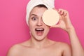 Studio shot of smiling woman with cosmetic sponge covering one eye,wrapped with white towel, female having perfect skin, standing Royalty Free Stock Photo