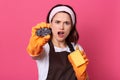 Studio shot of stressed sad woman standing isolated over pink wall with dirty sponge, being shocked, has astonished facial