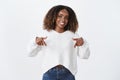 Studio shot of stylish african-american joyful female coworker in sweater pointing down and showing awesome promotion