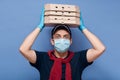 Studio shot of surprised delivery man dresses casual outfit, mask and latex gloves, holding stack of pizza`s boxes above his head
