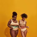 Studio shot of two african american women with different body weight measuring their waist using metric tape measure