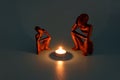 two wooden figurines sitting by a glowing fire of a candle like it was campfire on blue background