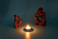 two wooden figurines sitting by a glowing fire of a candle like it was campfire on blue background