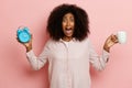 Studio shot of upset brunette girl in pink pajama with wide opened mouth, keeps alarm clock and cup, looks displeased Royalty Free Stock Photo