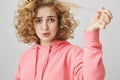 Studio shot of upset questioned curly-haired blonde woman pulling hair strand that remained straight after frizzle