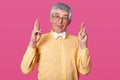 Studio shot of white haired senior raises hands up and crosses fingers. Elderly man wears yellow shirt poses in studio isolated Royalty Free Stock Photo