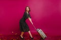 Studio shot of worried woman in black dress holding suitcase. Curly girl wears vintage dress afraids to be late and