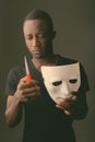 Studio shot of young black African man holding mask and scissors in dark room Royalty Free Stock Photo