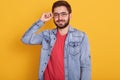 Studio shot of young attractive handsome man wearing red t shirt and denim jacket standing against yellow wall and smiling, keeps Royalty Free Stock Photo