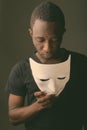 Studio shot of young black African man in dark room holding mask and looking down Royalty Free Stock Photo