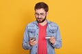 Studio shot of young european bearded man holding credit card and smartphone, standing with calm facial expression over yellow