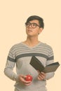 Studio shot of young handsome Asian man holding book and red apple while thinking Royalty Free Stock Photo