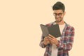 Studio shot of young happy Persian man smiling while wearing eyeglasses and reading book Royalty Free Stock Photo