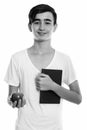 Studio shot of young happy Persian teenage boy smiling while holding book and red apple Royalty Free Stock Photo