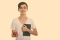 Studio shot of young happy Persian teenage boy smiling while holding book and red apple Royalty Free Stock Photo