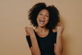 Young joyful excited dark skinned woman keeping fists clenched while posing against beige background