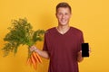 Studio shot of young man holding bunch of carrots and smart phone with blanl screen isolated over yellow background, male has Royalty Free Stock Photo