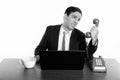 Studio shot of young stressed Persian businessman looking at old telephone with laptop and coffee cup on wooden table Royalty Free Stock Photo