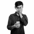 Studio shot of young stressed Persian teenage boy using mobile phone while covering face Royalty Free Stock Photo