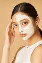 Studio shot of young woman with facial mask applied on half of her face looking at camera, receiving spa treatments